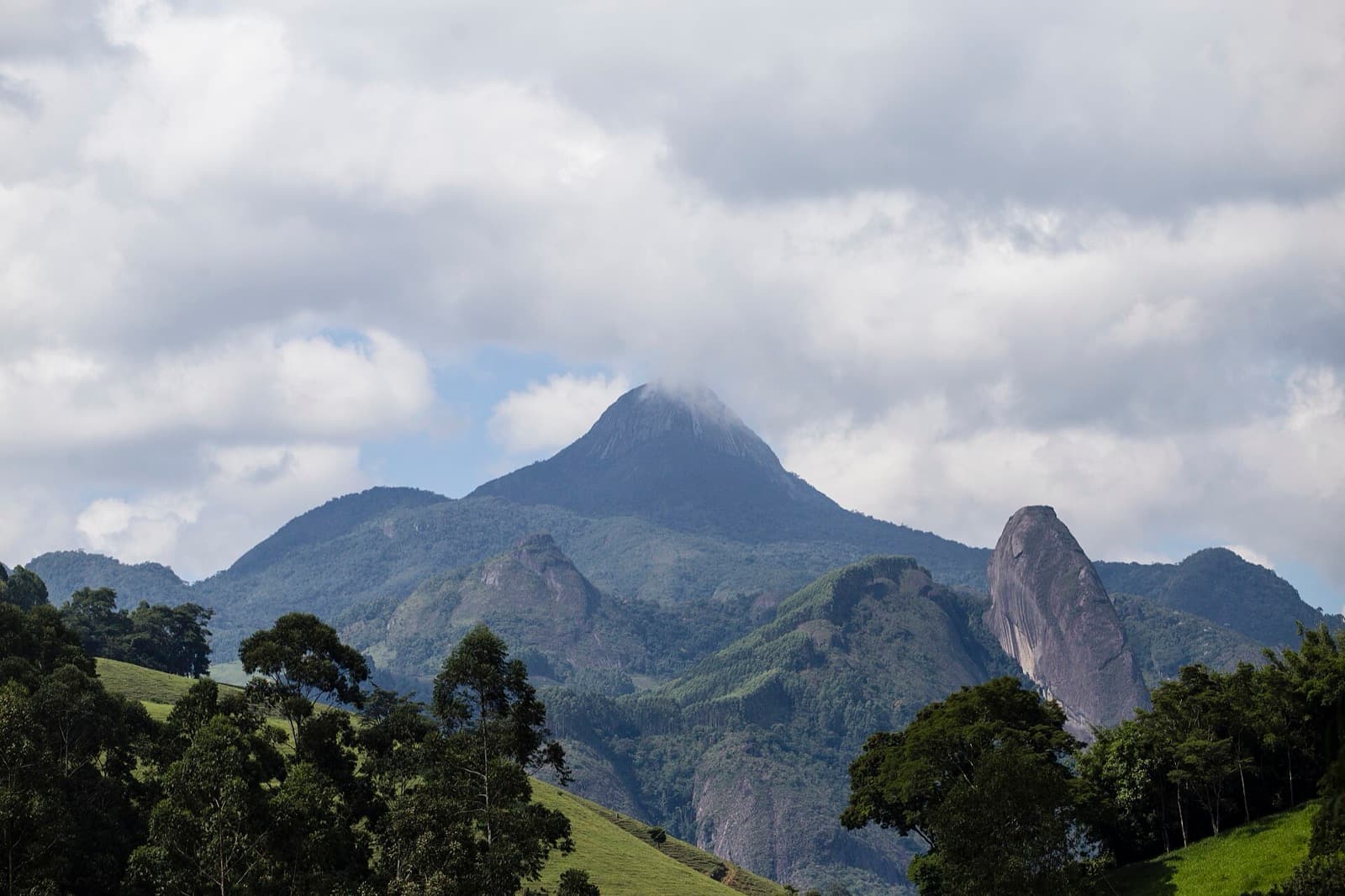 Pico do Forno Grande com o cume encoberto por nuvens, visto dos vales de Castelo, com um paredão de granito à direita e mata em primeiro plano