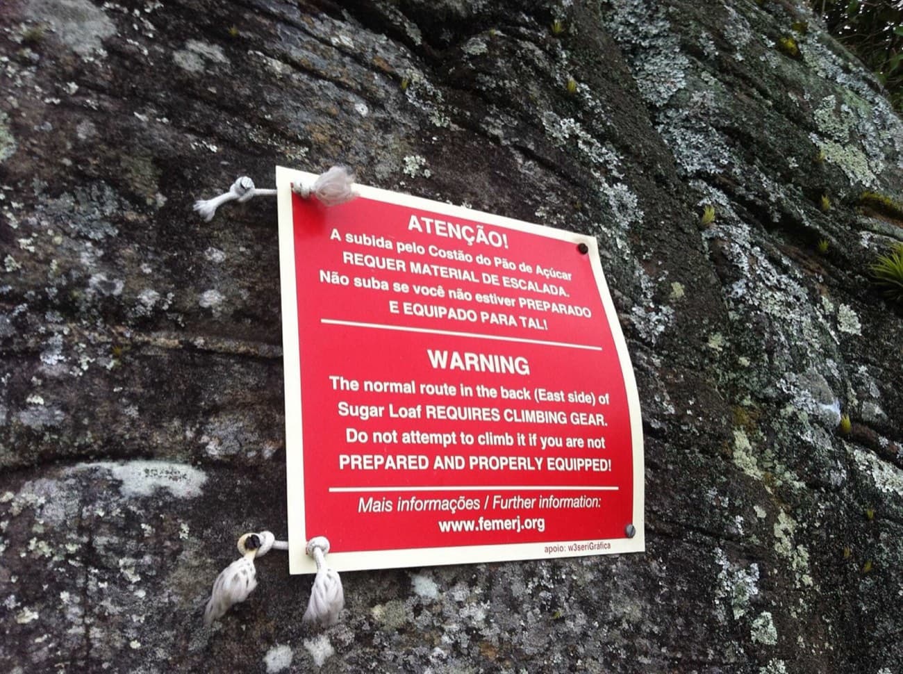 Placa vermelha da FEMERJ presa à rocha avisando que a subida pelo Costão exige material de escalada