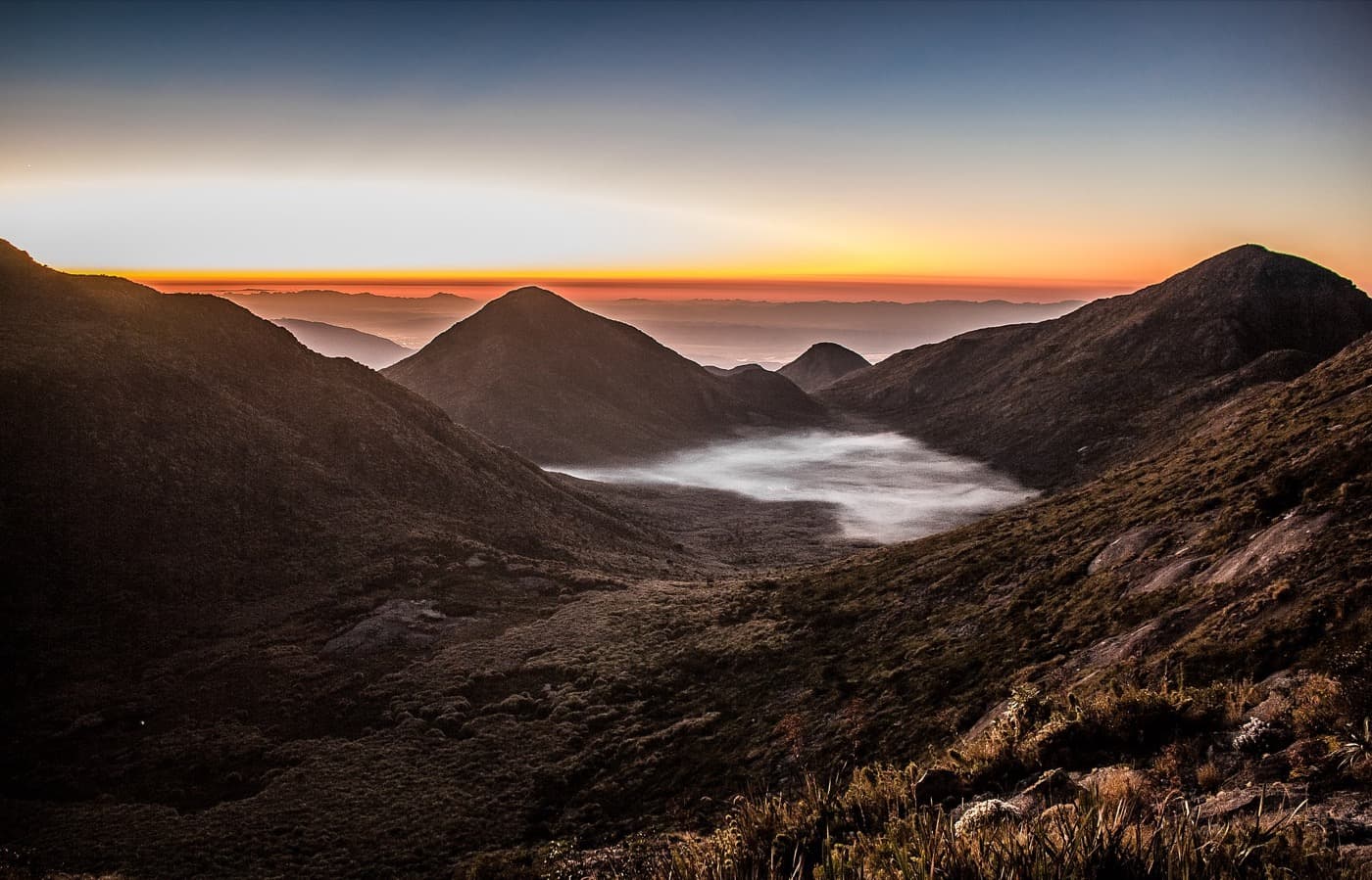 Amanhecer sobre o Vale do Ruah, na Serra Fina, com névoa no fundo do vale e picos escuros contra o horizonte alaranjado
