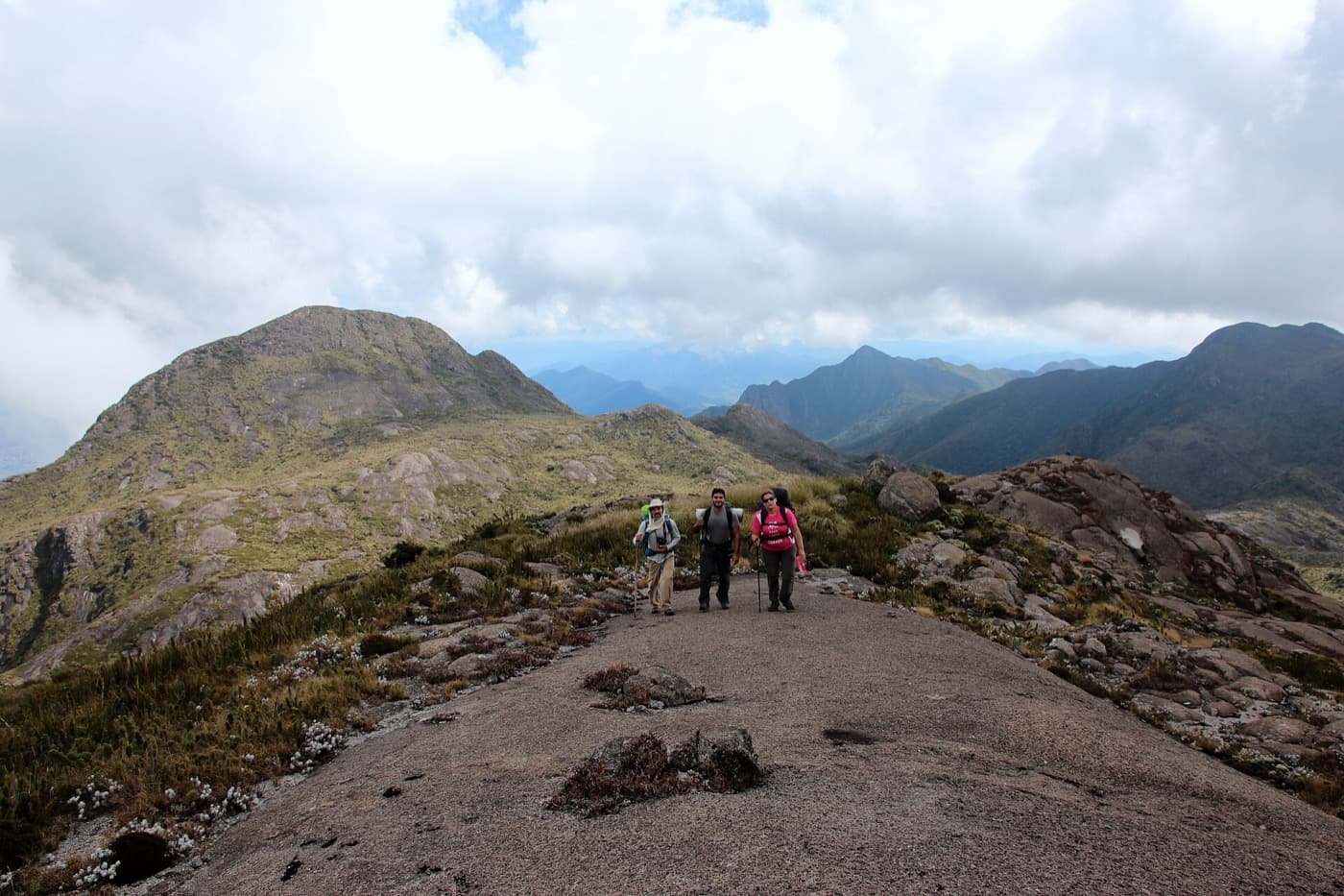Três montanhistas caminhando sobre a laje na subida da Pedra da Mina, com o Morro do Tartarugão ao fundo