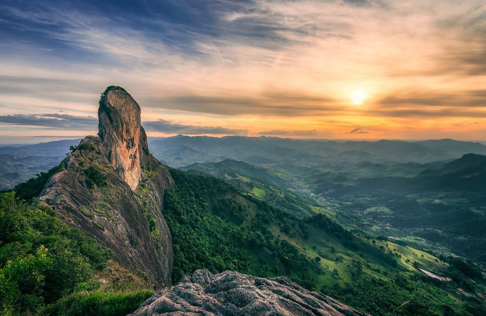 Perfil estreito da Pedra do Baú contra o pôr do sol, com vales e montanhas da Serra da Mantiqueira ao fundo