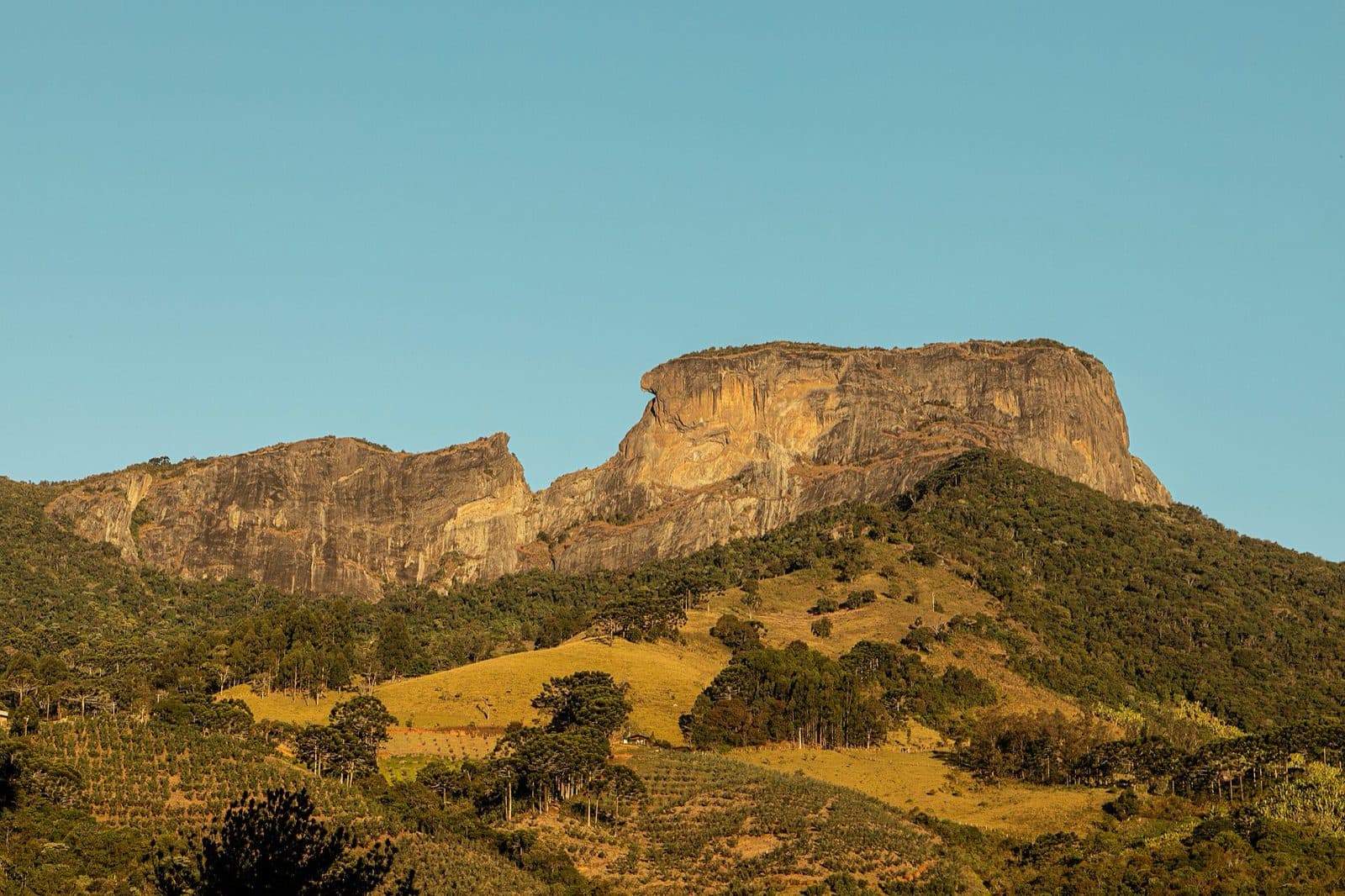 Bloco da Pedra do Baú iluminado pelo sol da tarde, acima de pastos e mata da encosta