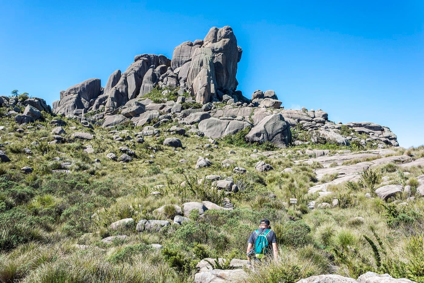 Trilheiro caminhando pelos campos de altitude em direção aos blocos de rocha empilhados das Prateleiras, sob céu azul