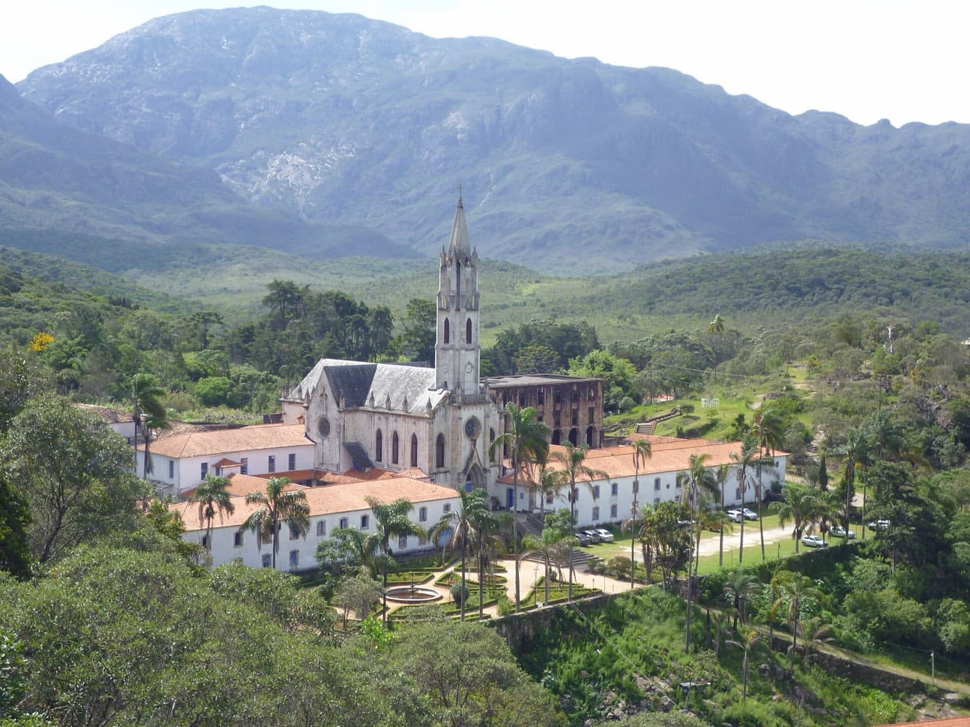 Igreja neogótica e prédios do Santuário do Caraça entre palmeiras, com a Serra do Caraça ao fundo
