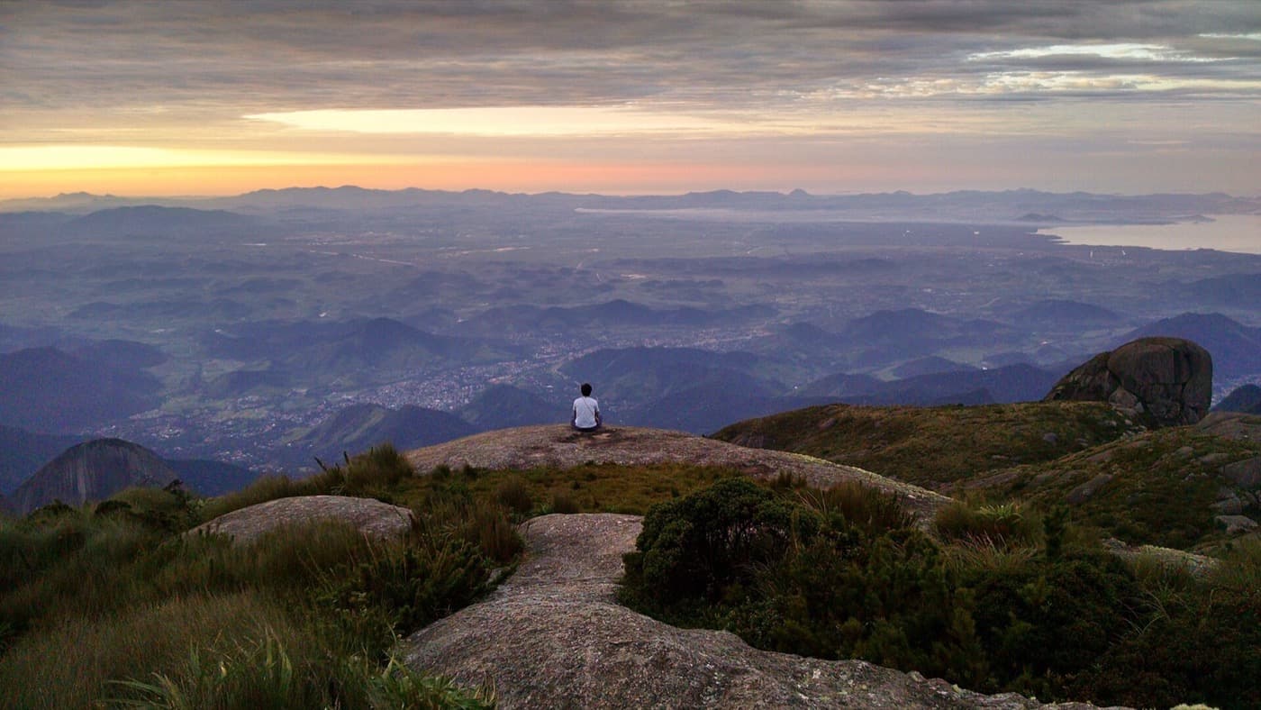 Pessoa sentada no alto da Pedra do Sino olhando a Baixada Fluminense e a Baía de Guanabara ao entardecer