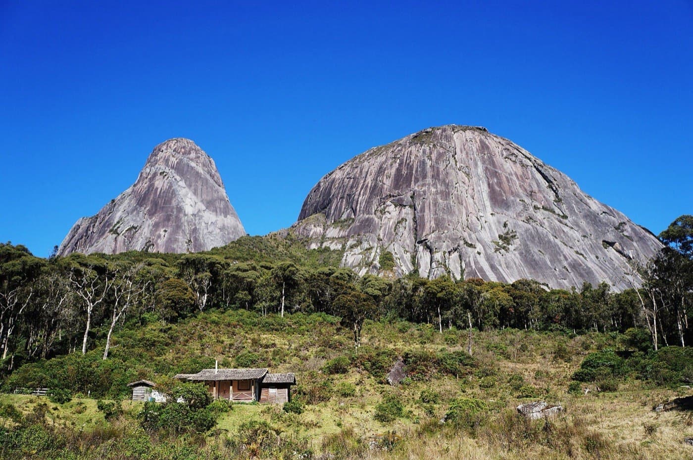 Pico Maior e Capacete, dois paredões de granito acima da mata, com uma casa de madeira no campo em primeiro plano