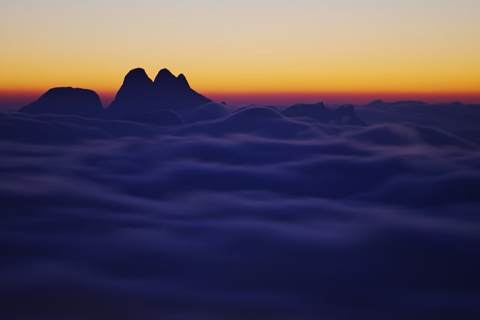 Silhueta dos Três Picos acima de um mar de nuvens ao amanhecer, vista de longe a partir da Pedra do Sino