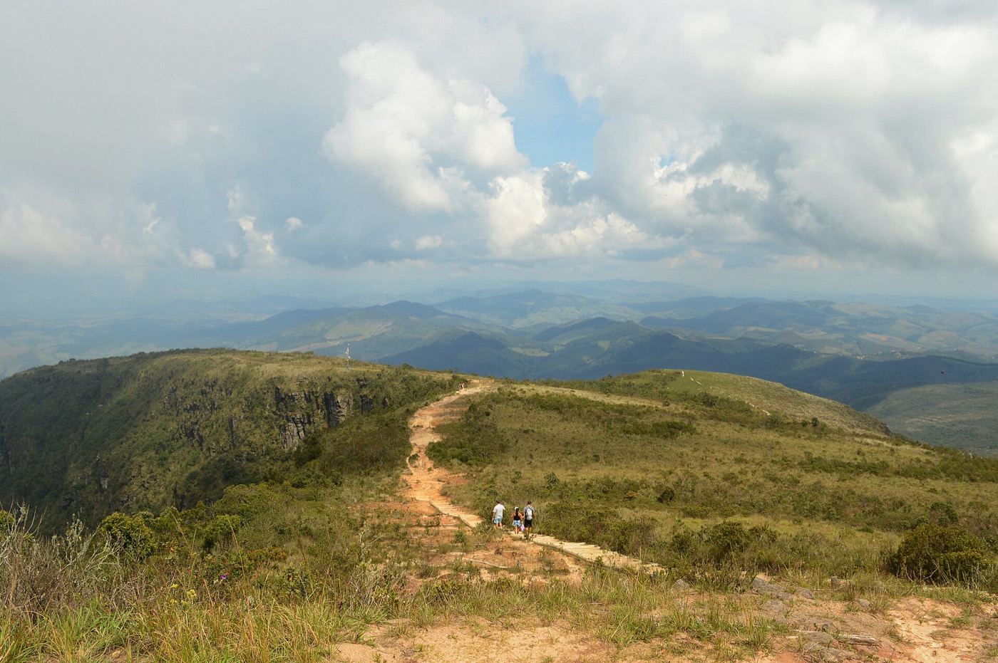 Trilha de terra na crista do Pico do Pião, com caminhantes e serras sob nuvens ao fundo