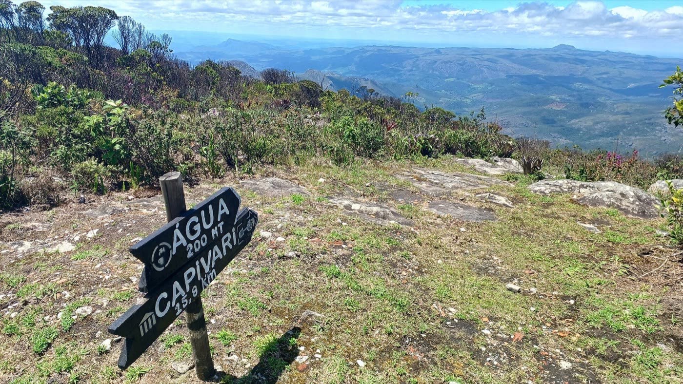 Placa de madeira perto do cume do Pico do Itambé indicando água a 200 m e Capivari a 15,9 km, com a serra ao fundo