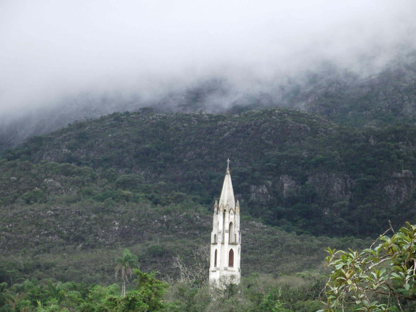 Torre da igreja do Caraça diante da serra coberta de neblina em dia nublado